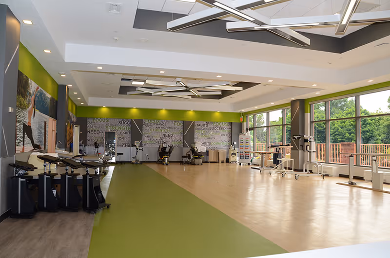 A spacious rehabilitation and exercise room with large windows letting in natural light. The room features various exercise equipment including stationary bikes and other rehabilitation machines. The walls have motivational words and images of people exercising. The floor has a green and wood-tone design, and modern ceiling lights are installed.