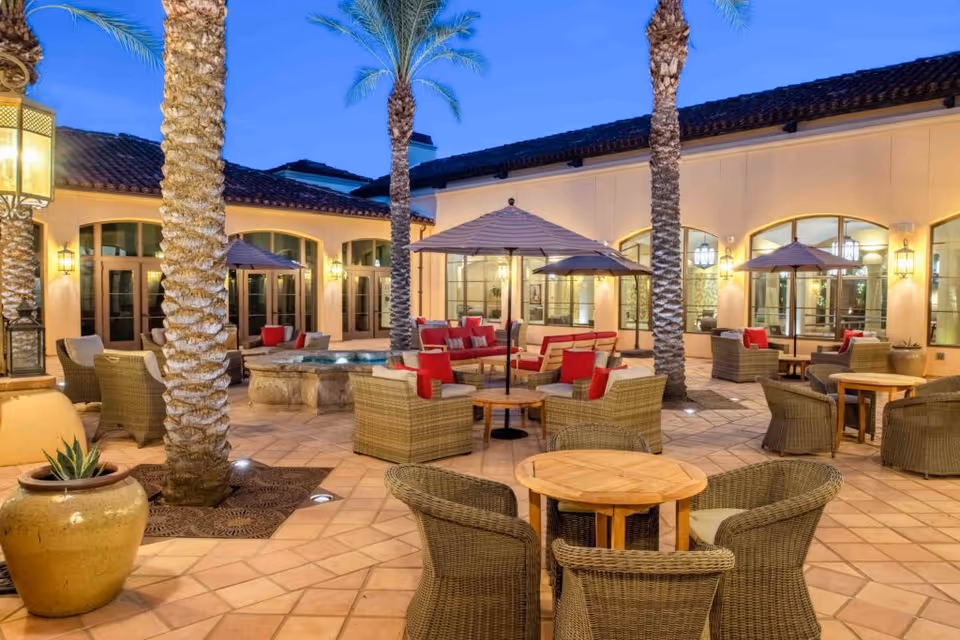 Outdoor patio area at Maravilla Scottsdale with wicker chairs and tables, red cushions, umbrellas, palm trees, and warm lighting against a twilight sky.
