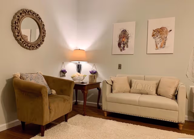 A cozy living room with a beige sofa adorned with pillows, including one that says 'Home', a brown armchair with a decorative pillow, a wooden side table with a lamp and small purple flowers, a round ornate mirror on the wall, and two framed pictures of a bear and a tiger above the sofa.
