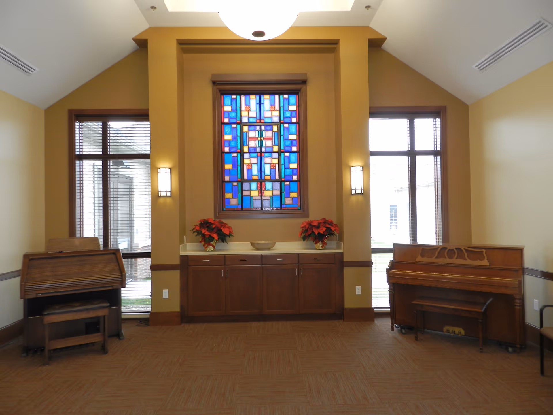 Interior room with a high ceiling featuring a colorful stained glass window centered above a wooden cabinet with two poinsettia plants. On either side of the cabinet are large windows with blinds. To the left is an organ with a matching wooden bench, and to the right is a piano with a bench. The walls are painted a warm beige color and the floor is carpeted.