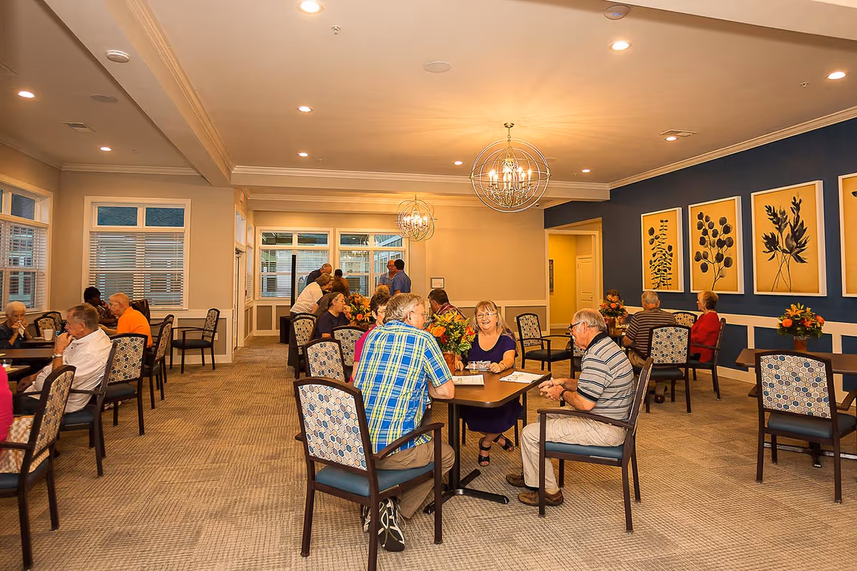 A dining room in a senior living facility with several elderly people sitting at tables, chatting and enjoying their time. The room has patterned chairs, beige carpeting, and blue accent walls decorated with framed botanical prints. Modern spherical chandeliers hang from the ceiling, and there are flower arrangements on the tables.
