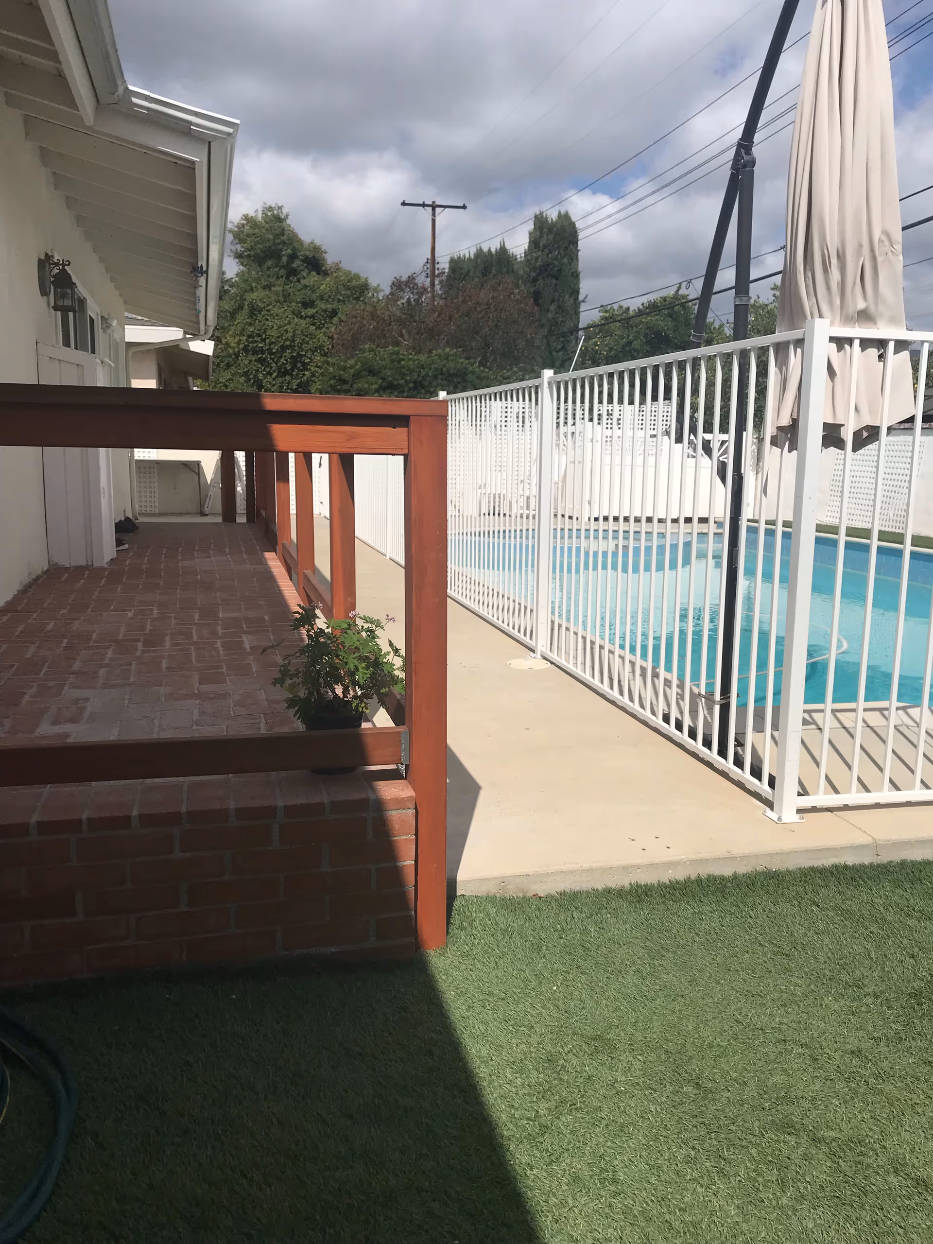 Outdoor area of a senior living facility showing a fenced swimming pool with a closed umbrella, a brick patio with a wooden railing, some greenery, and a partly cloudy sky.