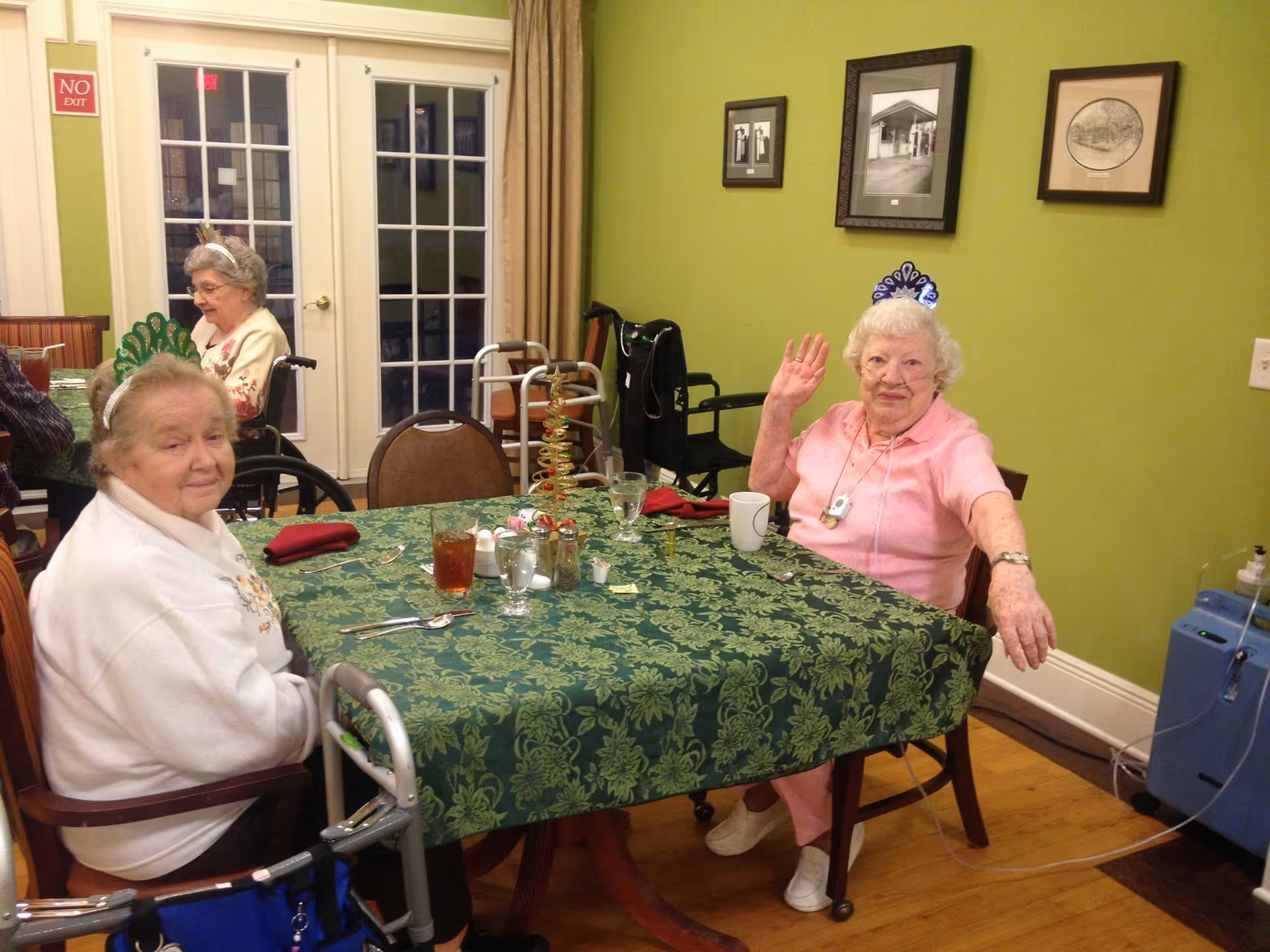 Two elderly women sitting at a dining table with a green patterned tablecloth in a room with green walls. One woman is wearing a white sweater and a green tiara, and the other is wearing a pink outfit and a blue tiara, waving at the camera. There are glasses, cups, and utensils on the table. In the background, another elderly woman is seated near glass doors. A walker and a wheelchair are visible, along with framed pictures on the wall.