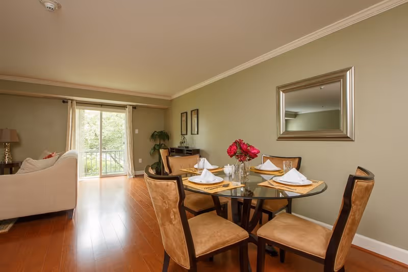 A dining area with a round glass table set for four with white napkins and plates, surrounded by four cushioned chairs. The room has wooden flooring, a large mirror on the wall, and a sliding glass door with curtains leading to an outdoor space. A beige sofa and a side table with a lamp are visible in the adjacent living area.