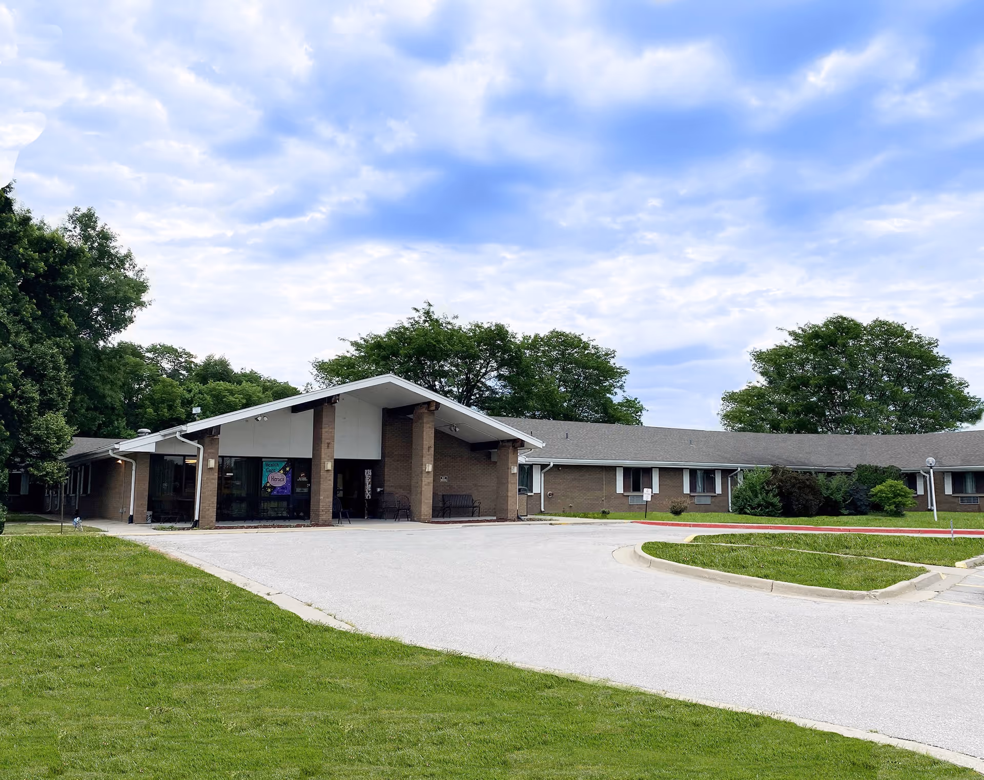 Exterior view of a single-story brick building with a covered entrance, surrounded by green grass and trees under a partly cloudy sky.