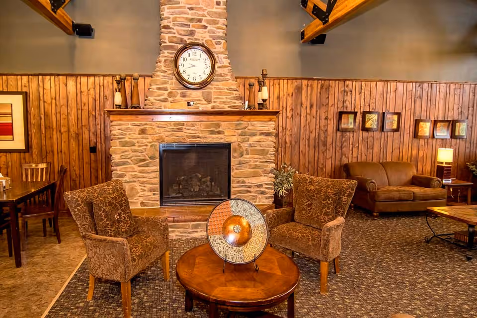 Cozy living room area with a stone fireplace centered on a wood-paneled wall. Above the fireplace is a round clock. In front of the fireplace are two patterned armchairs and a round wooden coffee table with a decorative plate. To the right is a brown sofa with a side table and lamp, and framed pictures hang on the wall above it. To the left, a dining table with chairs is partially visible.