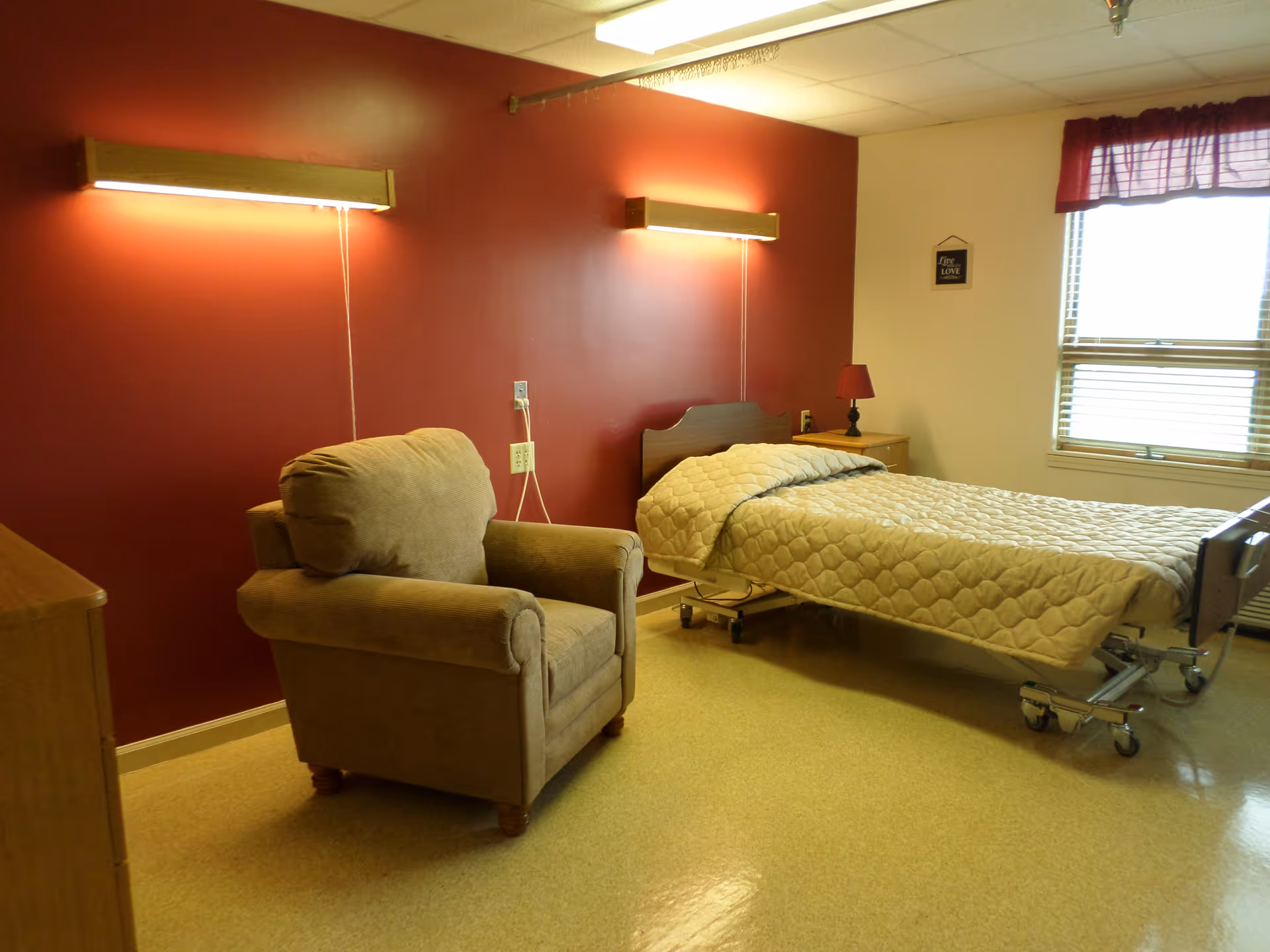 A simple, clean bedroom in a healthcare facility with a single hospital-style bed covered with a beige quilt, a cushioned armchair, a wooden nightstand with a small red lamp, and a window with blinds and a red valance. The walls are painted red and white, with two wall-mounted lights above the bed.