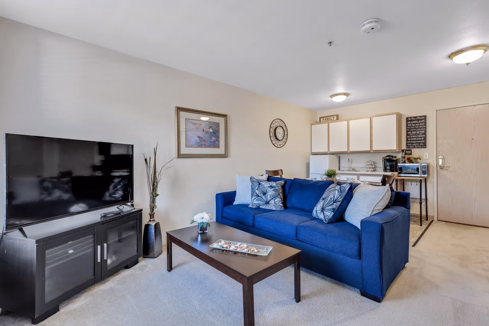 A cozy living room area with a blue sofa adorned with patterned and plain cushions, a dark wooden coffee table with a decorative tray and small flower vase, a flat-screen TV on a black TV stand, and a small kitchenette in the background with white cabinets, a mini fridge, microwave, and coffee maker. The walls are light-colored with a framed picture, a clock, and decorative signs.
