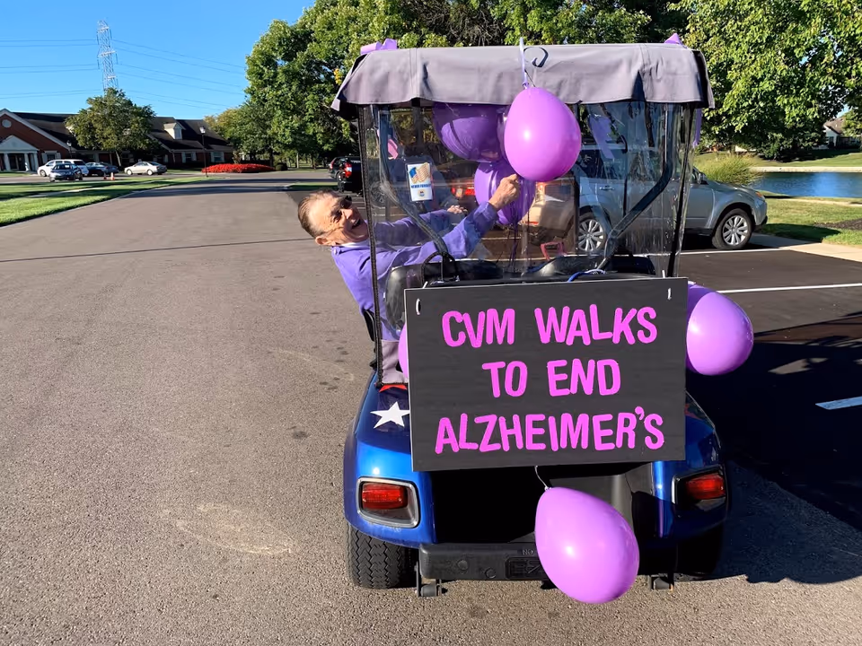 A person sitting in a blue golf cart decorated with purple balloons and a sign that reads 'CVM WALKS TO END ALZHEIMER'S' in pink letters. The person is smiling and holding one of the balloons. The setting is an outdoor parking area with trees, cars, and buildings in the background.