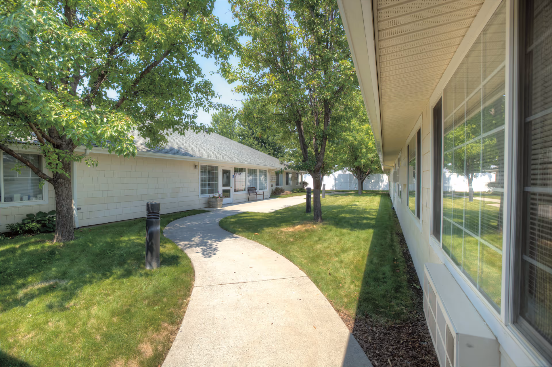 A curved concrete pathway runs through a grassy courtyard area with several trees and bushes. The pathway is flanked by single-story buildings with large windows, and there is a bench near one of the building entrances. The scene is bright and sunny with clear blue skies.