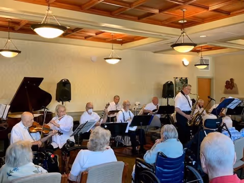 A group of elderly musicians performing indoors with various instruments including a violin, saxophone, and guitar. Several elderly audience members are seated and watching the performance in a room with a wooden coffered ceiling and beige walls.