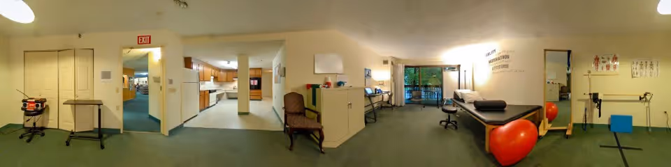 A wide view of a senior living facility's interior showing a physical therapy or rehabilitation area with exercise equipment including a large red exercise ball, a treatment table, chairs, and anatomical charts on the walls. There is a kitchen area visible in the background and a glass door leading to an outdoor space.