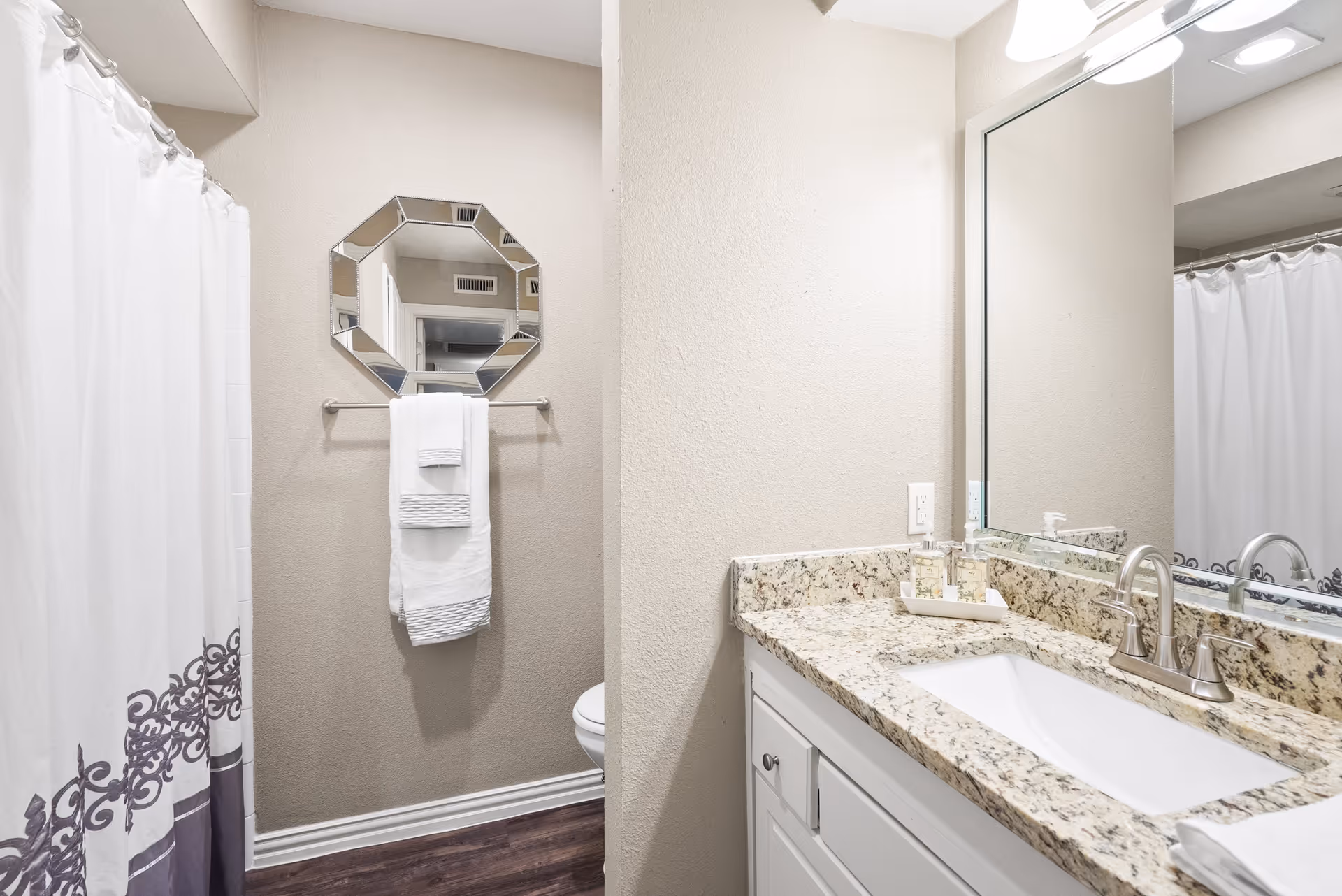 A clean bathroom with a granite countertop and sink, large mirror, towel on a rack, and a shower with a patterned curtain.