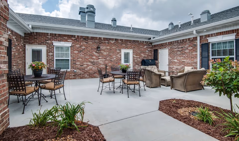 Outdoor patio area with brick walls, featuring two round tables with chairs and flower pots, a seating area with cushioned wicker furniture, and some landscaped plants along the edges.