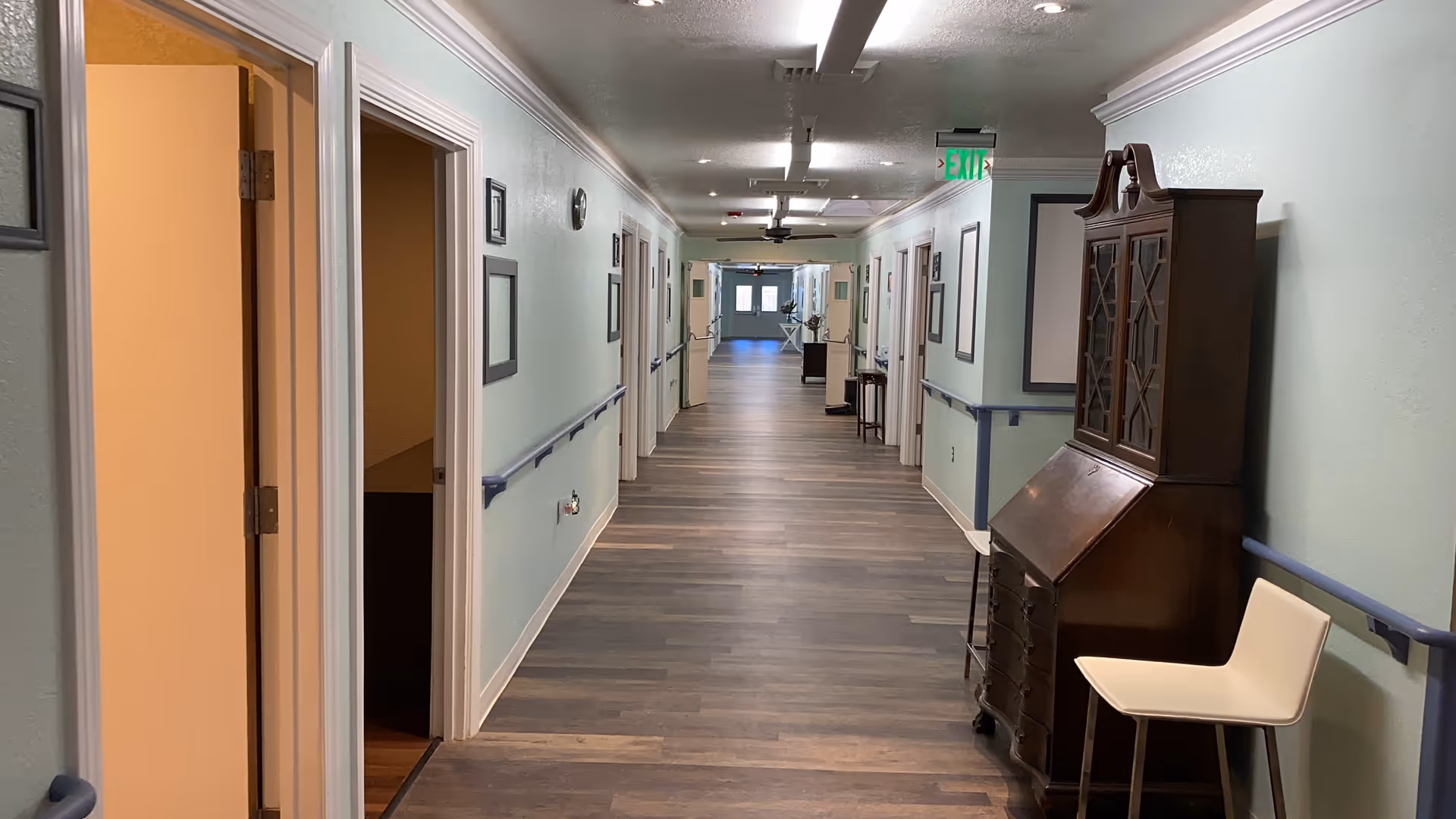 Interior hallway of a senior living facility with doors on both sides, wall handrails, and a wooden cabinet and chair in the foreground.