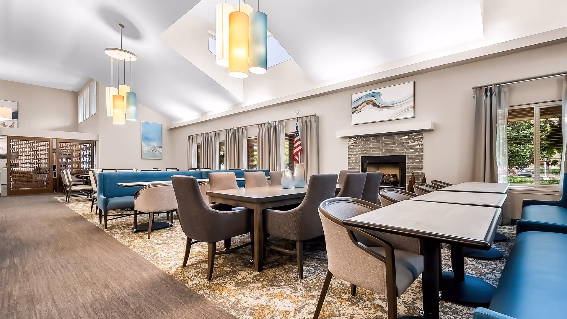 A spacious and well-lit dining room with multiple tables and chairs arranged neatly. The room features a high vaulted ceiling with modern pendant lights, large windows with curtains, a fireplace with artwork above it, and an American flag near the window. The decor includes a mix of blue and beige tones with patterned carpeting.