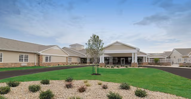Exterior view of a single-story senior living facility with beige siding and stone accents, a covered entrance with white columns, a green lawn, and landscaped areas with small shrubs and rocks under a partly cloudy sky.