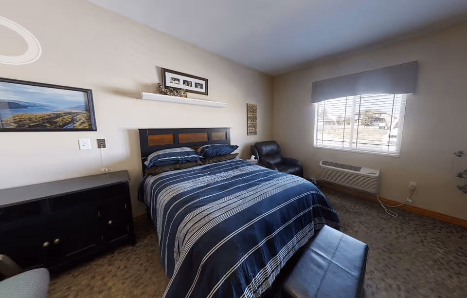A bedroom with a bed covered in a blue and white striped comforter, two pillows, a black headboard, a black armchair in the corner, a window with blinds and a gray valance, a black cabinet, and framed pictures on the beige walls.