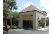 Entrance area of The Meadows At Cypress Gardens facility with a covered driveway, surrounded by palm trees and greenery under a partly cloudy sky.