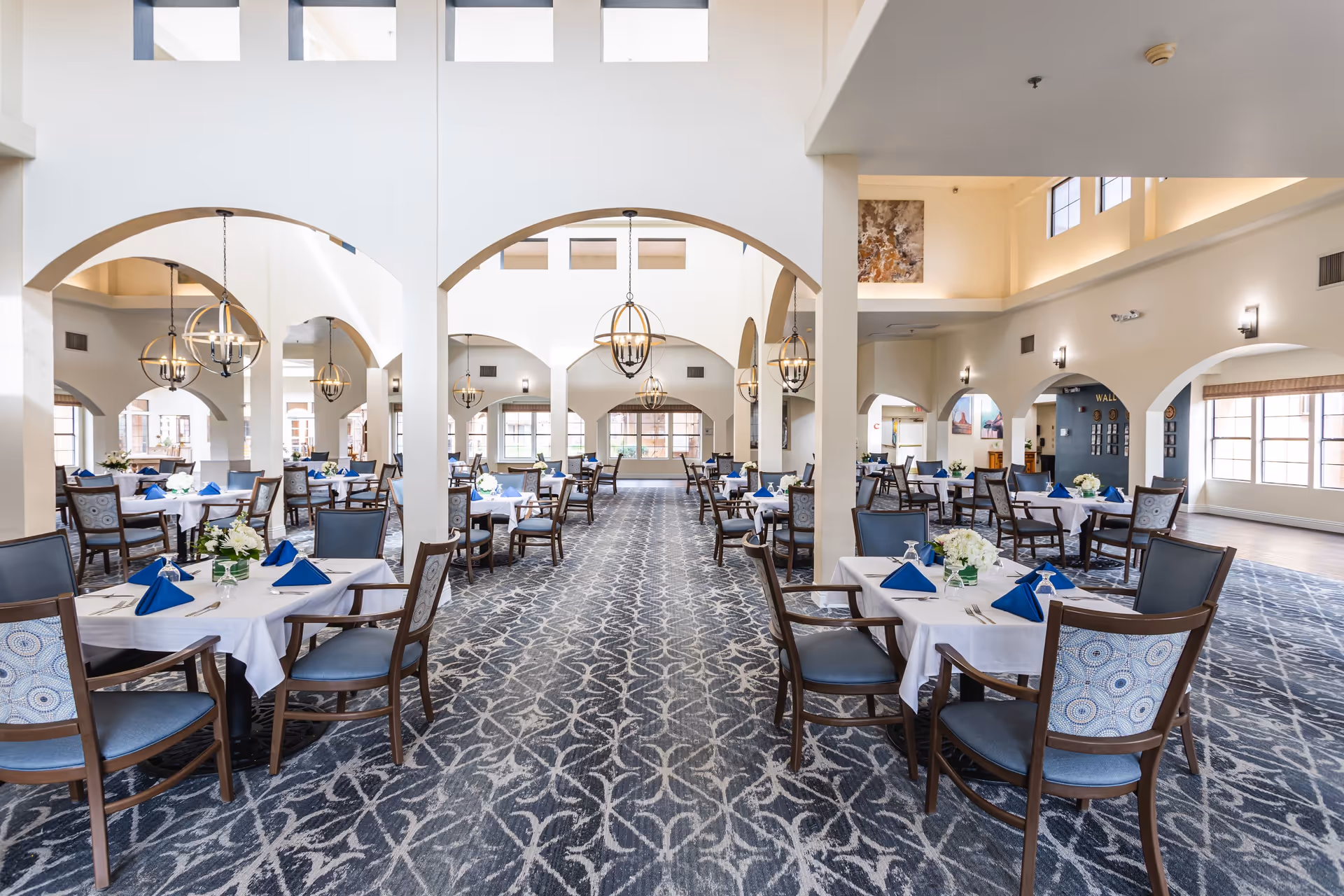 Spacious dining room with neatly set tables, blue napkins, chandeliers, and arched columns.