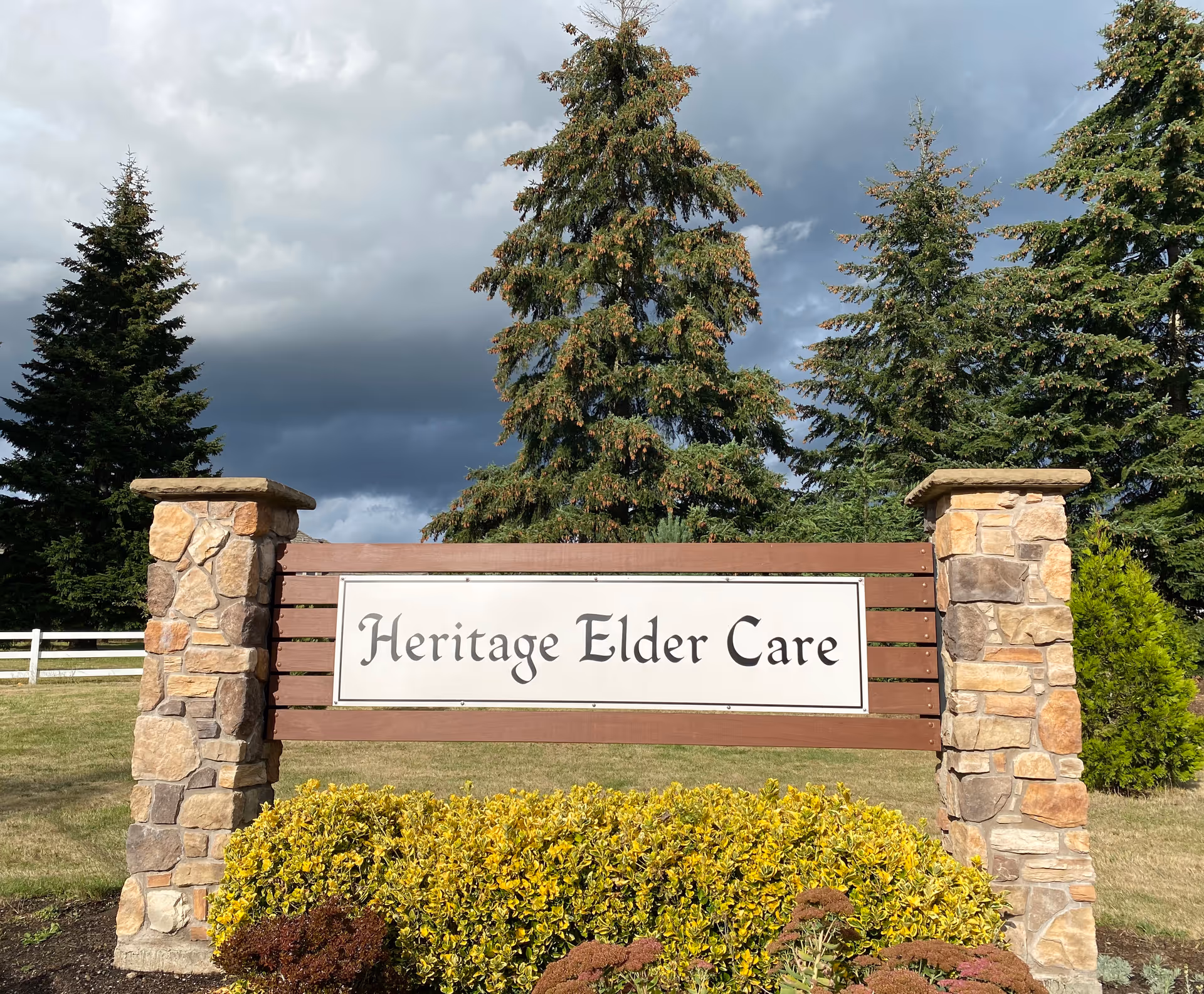 A large outdoor sign for Heritage Elder Care mounted on wooden slats supported by two stone pillars, with green bushes in front and tall evergreen trees in the background under a cloudy sky.