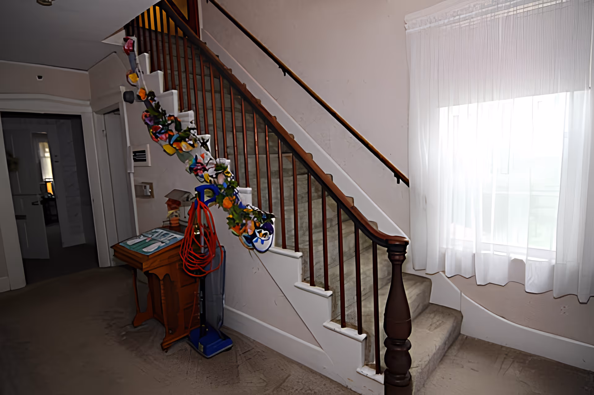 Interior view of a carpeted staircase with wooden handrails and spindles. The staircase is decorated with a colorful garland. Next to the staircase is a small wooden table with a green book or binder on top and a vacuum cleaner leaning against it. A large window with sheer white curtains allows natural light to brighten the space.