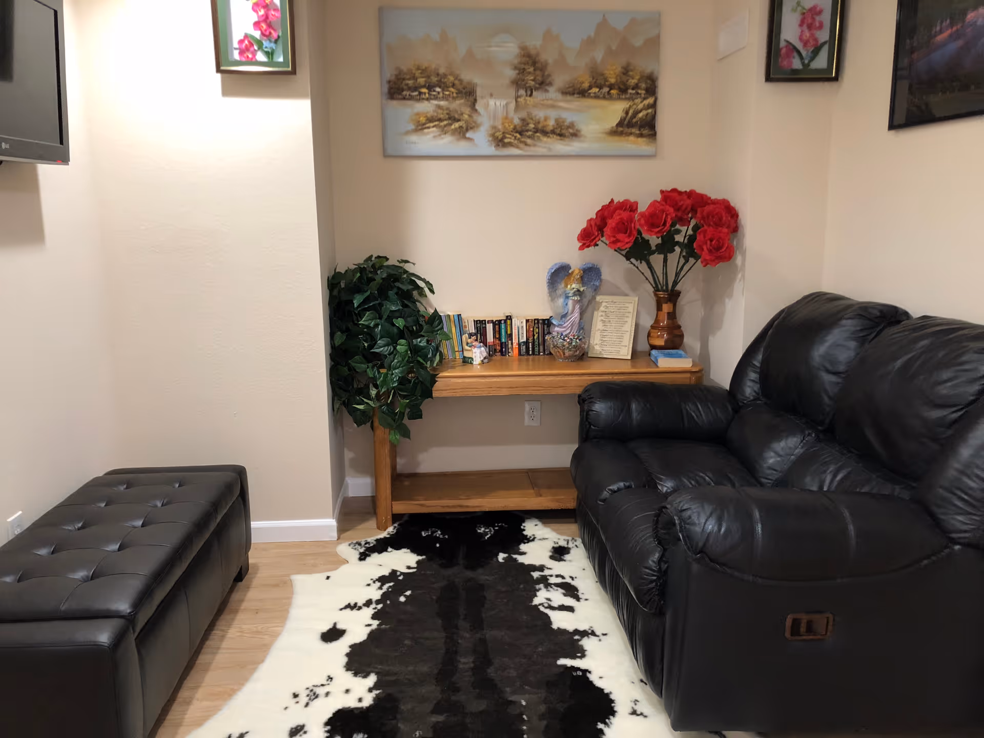 A cozy living room area with a black leather recliner sofa on the right and a black tufted bench on the left. A wooden console table against the back wall holds a collection of books, a decorative angel figurine, a vase with red flowers, and a framed document. Above the table hangs a landscape painting, and two smaller floral pictures are on the adjacent walls. A black and white cowhide rug is on the wooden floor.