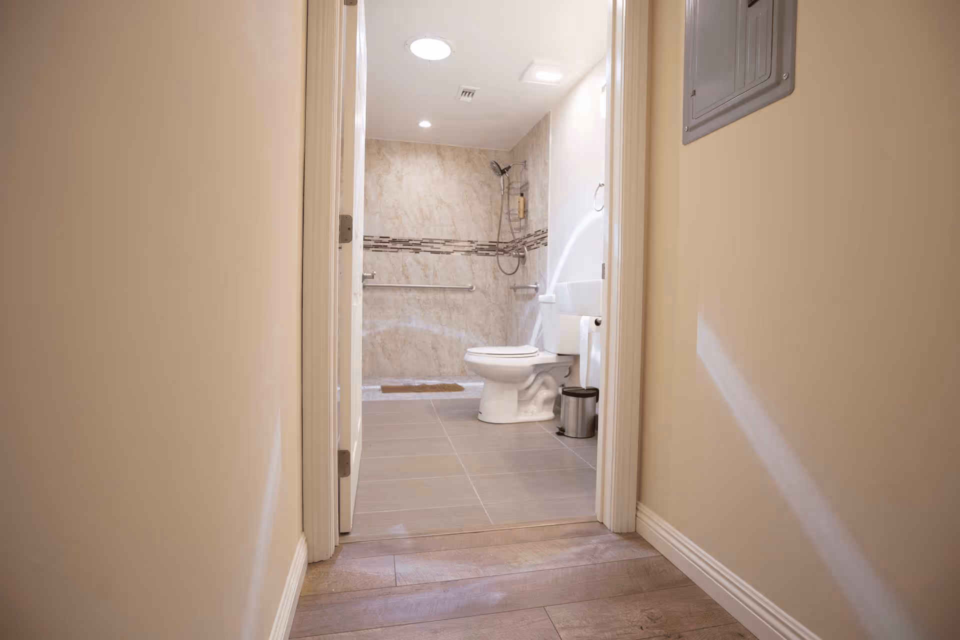 View down a hallway into a bathroom featuring a toilet, a small trash can, and a walk-in shower with a handheld showerhead and grab bars. The bathroom has beige tiled walls with a decorative horizontal strip and gray floor tiles.