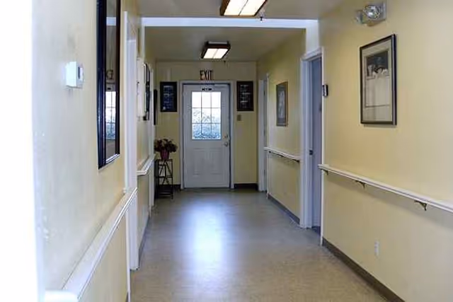 A clean, well-lit hallway with pale yellow walls, handrails on both sides, framed pictures hanging on the walls, and a door with a window at the end marked with an exit sign above it.