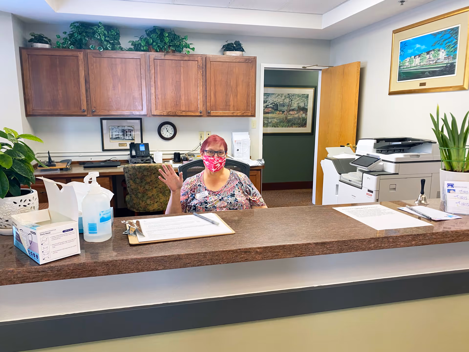 A woman wearing a red floral face mask and patterned shirt is sitting behind a reception desk in an office area. She is waving with her right hand. The desk has a box of face masks, a bottle of hand sanitizer, clipboards with papers, and a bell. Behind her are wooden cabinets, a clock, a phone, and framed pictures on the wall. To the right, there is a printer and a potted plant on the counter. The office door is open, showing a hallway with more framed artwork.