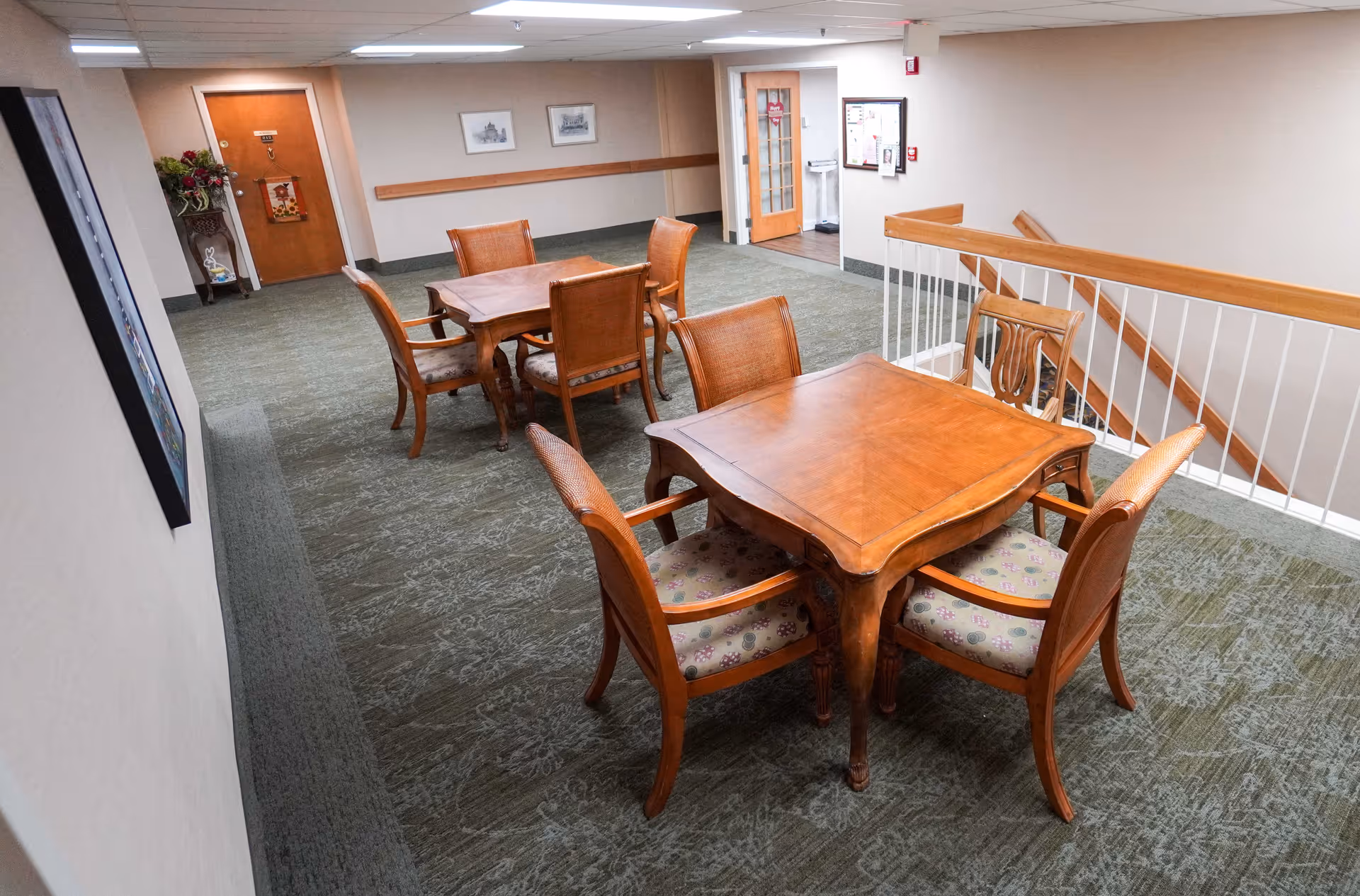Interior view of a common area in a senior living facility with two wooden tables, each surrounded by four wooden chairs with patterned cushions. The area has carpeted flooring, beige walls, framed pictures on the walls, and a staircase with a wooden handrail and white balusters. There is a door and a small table with flowers in the background.