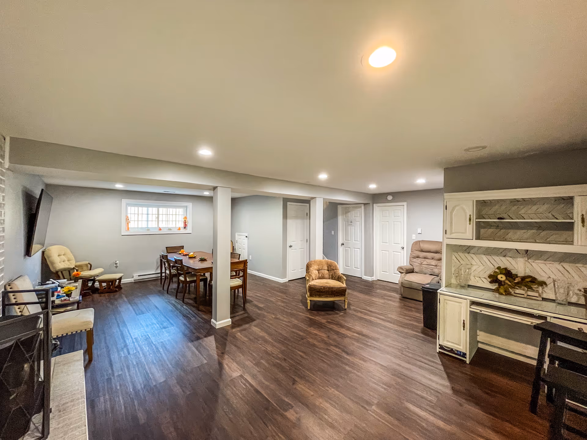 A spacious, well-lit common area with wooden flooring and light gray walls. The room features a dining table with chairs near a window, several armchairs, a wall-mounted TV, and a white cabinet with glass shelves. Recessed ceiling lights illuminate the space.