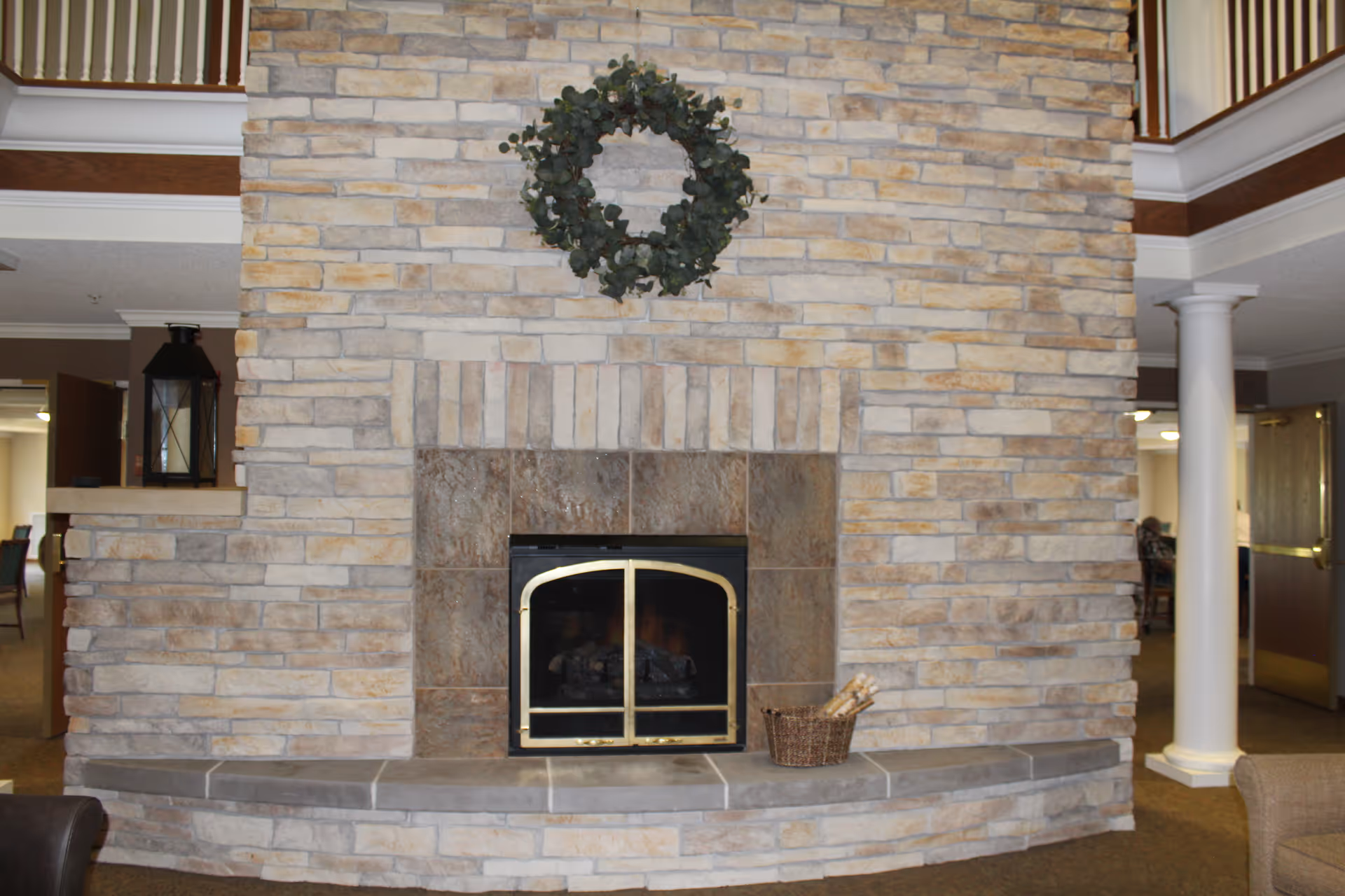 A stone fireplace with a metal framed glass door, a green wreath hanging above it, and a basket with firewood on the hearth. The fireplace is set in a spacious room with carpeted floor, white columns, and wooden railings on the upper level.