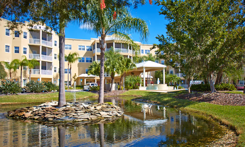 Exterior view of a multi-story senior living facility named Cypress Palms with balconies, surrounded by palm trees and other greenery. In the foreground, there is a pond with a small island of rocks and two palm trees, along with a water fountain. The sky is clear and blue.