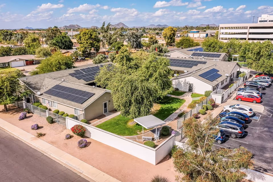Aerial view of a senior living facility with multiple single-story buildings featuring solar panels on the roofs. The facility has a fenced green courtyard with trees, shrubs, and a covered bench area. There is a parking lot with several cars parked, and the surrounding area includes other buildings and trees under a partly cloudy sky.