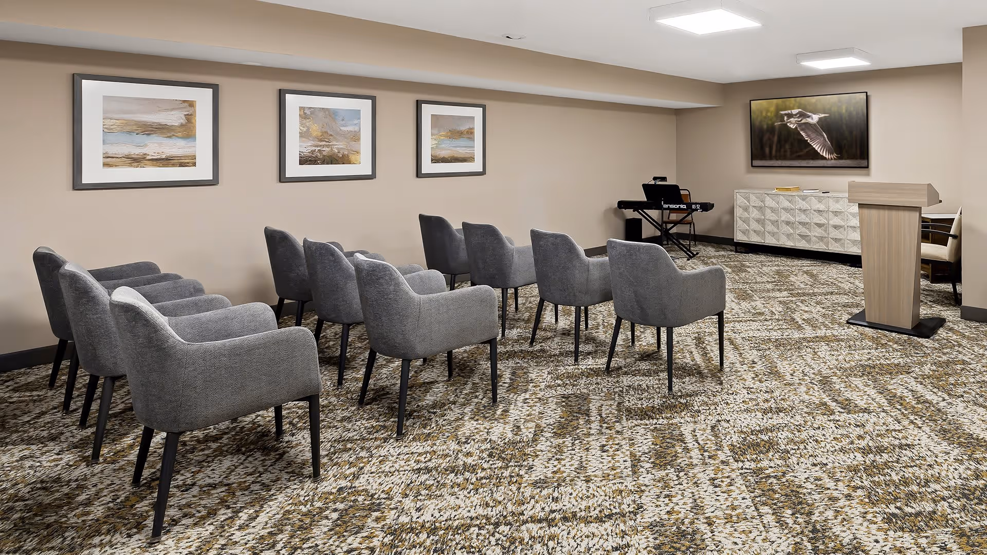 A small conference or presentation room with two rows of gray upholstered chairs facing a wooden podium and a large flat-screen TV mounted on the wall. The room has beige walls with three framed landscape paintings and patterned carpet flooring. A keyboard is placed in the corner near the TV.