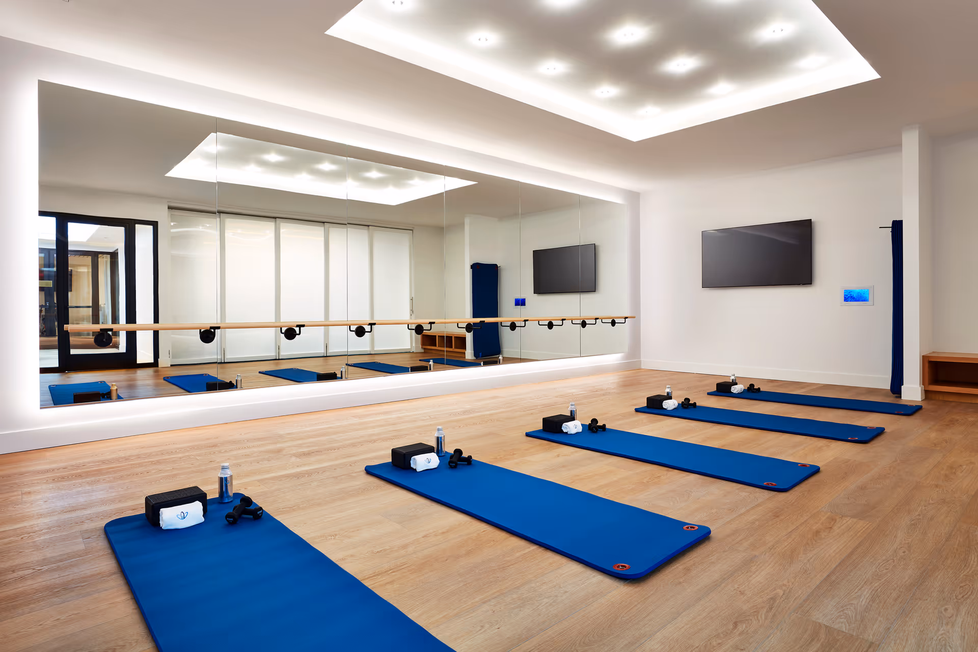 A bright exercise room with wooden flooring and a large wall mirror. Five blue exercise mats are laid out on the floor, each equipped with a water bottle, towel, yoga block, and small dumbbells. The room features a ballet barre along the mirrored wall and a flat-screen TV mounted on the opposite wall. The ceiling has recessed lighting, and there is a door leading to another area.