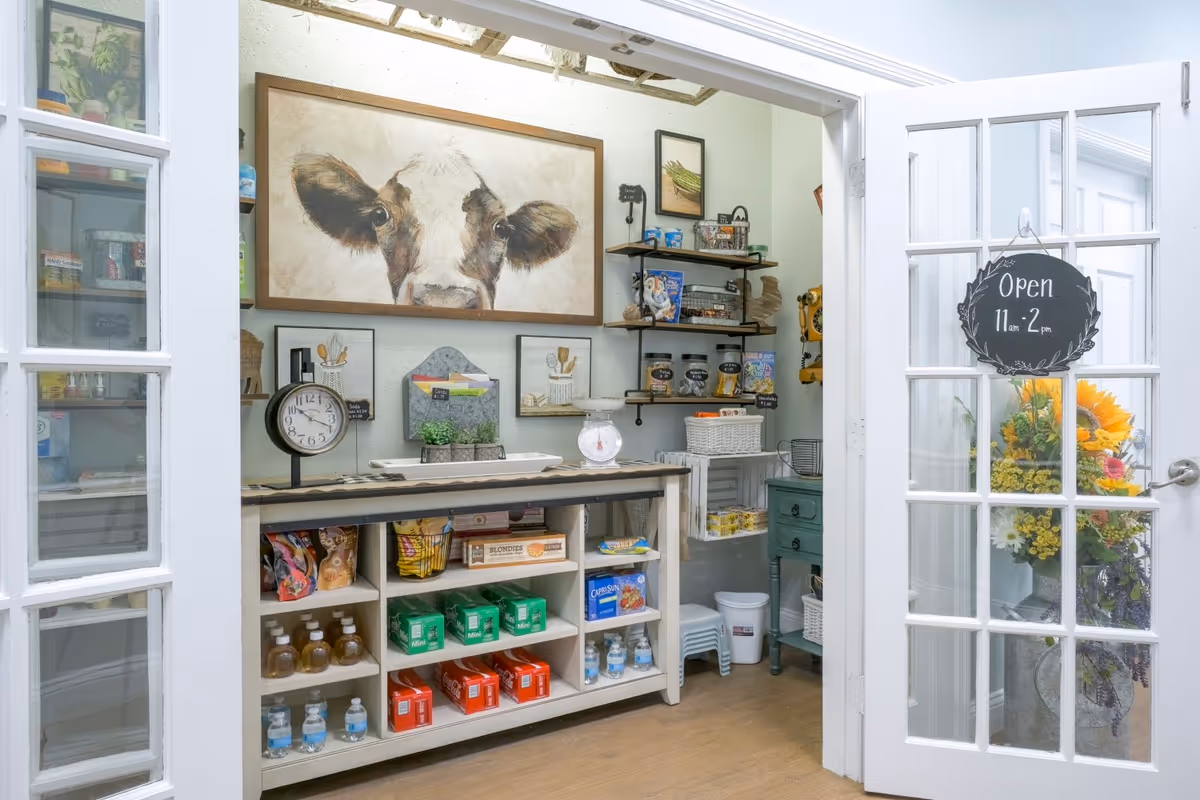 A small pantry or snack area with shelves stocked with various snacks, bottled water, and other items. There is a large painting of a cow on the wall above the counter. The room has white French doors, one of which has a sign that reads 'Open 11 am - 2 pm'. A vase with sunflowers and other flowers is visible near the door.