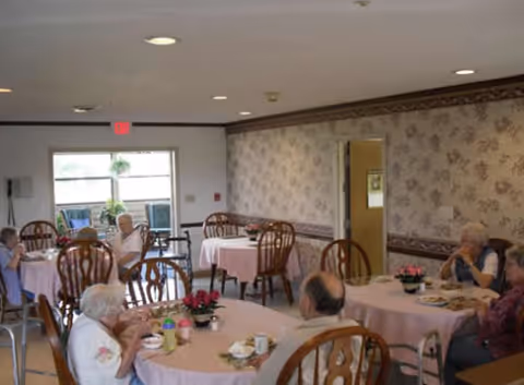 Residents sitting at round tables with pink tablecloths in a communal dining room, eating and talking.