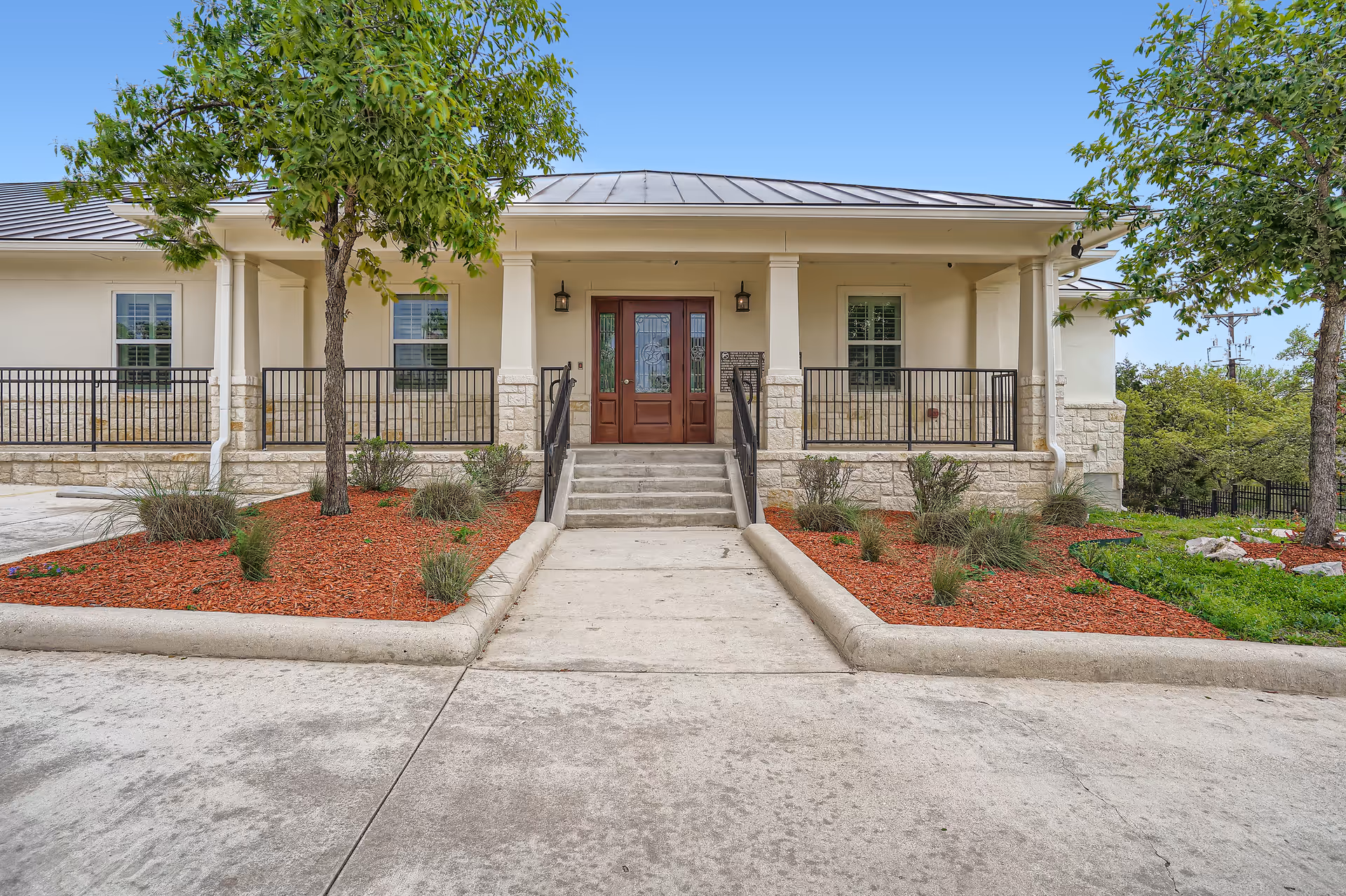 Front entrance of a senior living building with steps leading to double doors, a covered porch, railing, and landscaped beds with trees.
