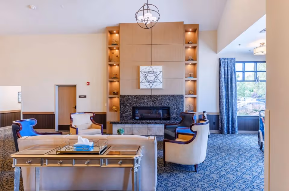 Bright, elegant senior living lounge with upholstered chairs and sofas arranged around a central fireplace and built-in shelving.