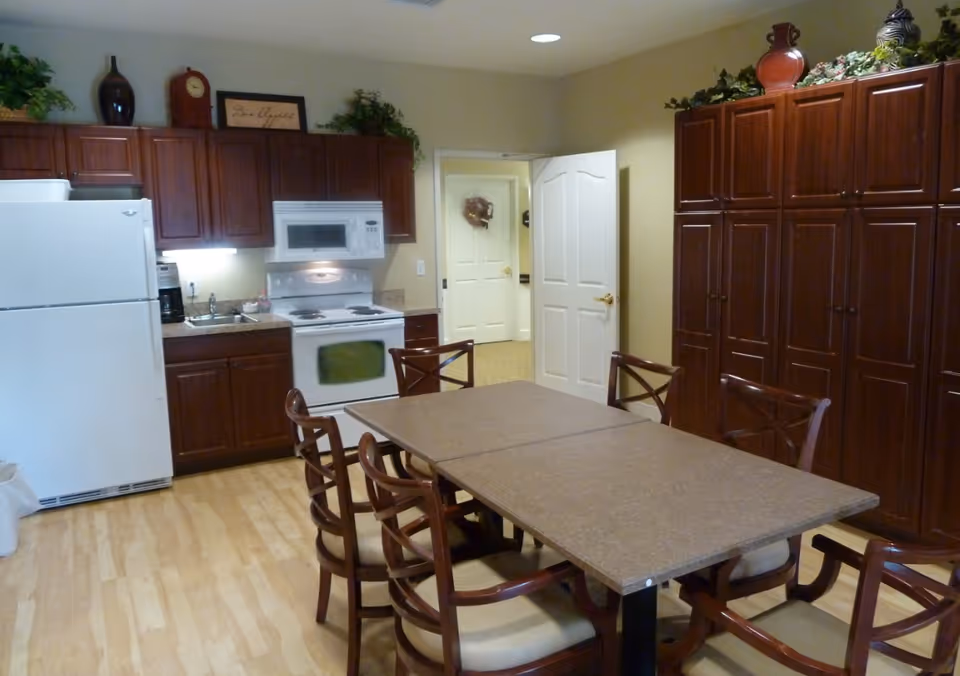 Interior view of a kitchen and dining area with wooden cabinets, a white refrigerator, stove, microwave, and a rectangular dining table surrounded by six wooden chairs with cushioned seats. The room has light-colored walls and flooring, with decorative plants and vases on top of the cabinets.