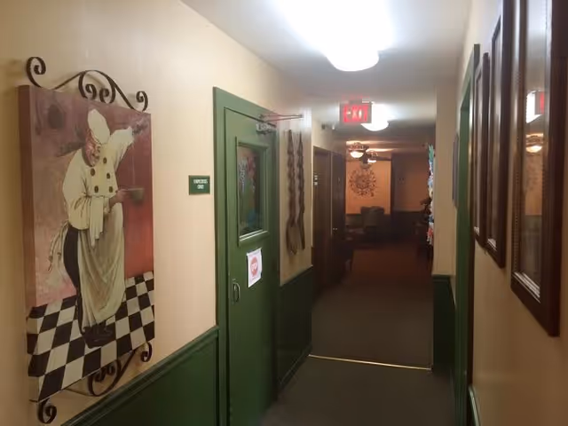 A hallway in Walnut Street Assisted Living with green lower walls and beige upper walls. On the left wall, there is a painting of a chef in a white uniform and hat. A green door with a sign reading 'Employees Only' is also visible. The hallway leads to a room with chairs and wall decorations, illuminated by ceiling lights and an exit sign.
