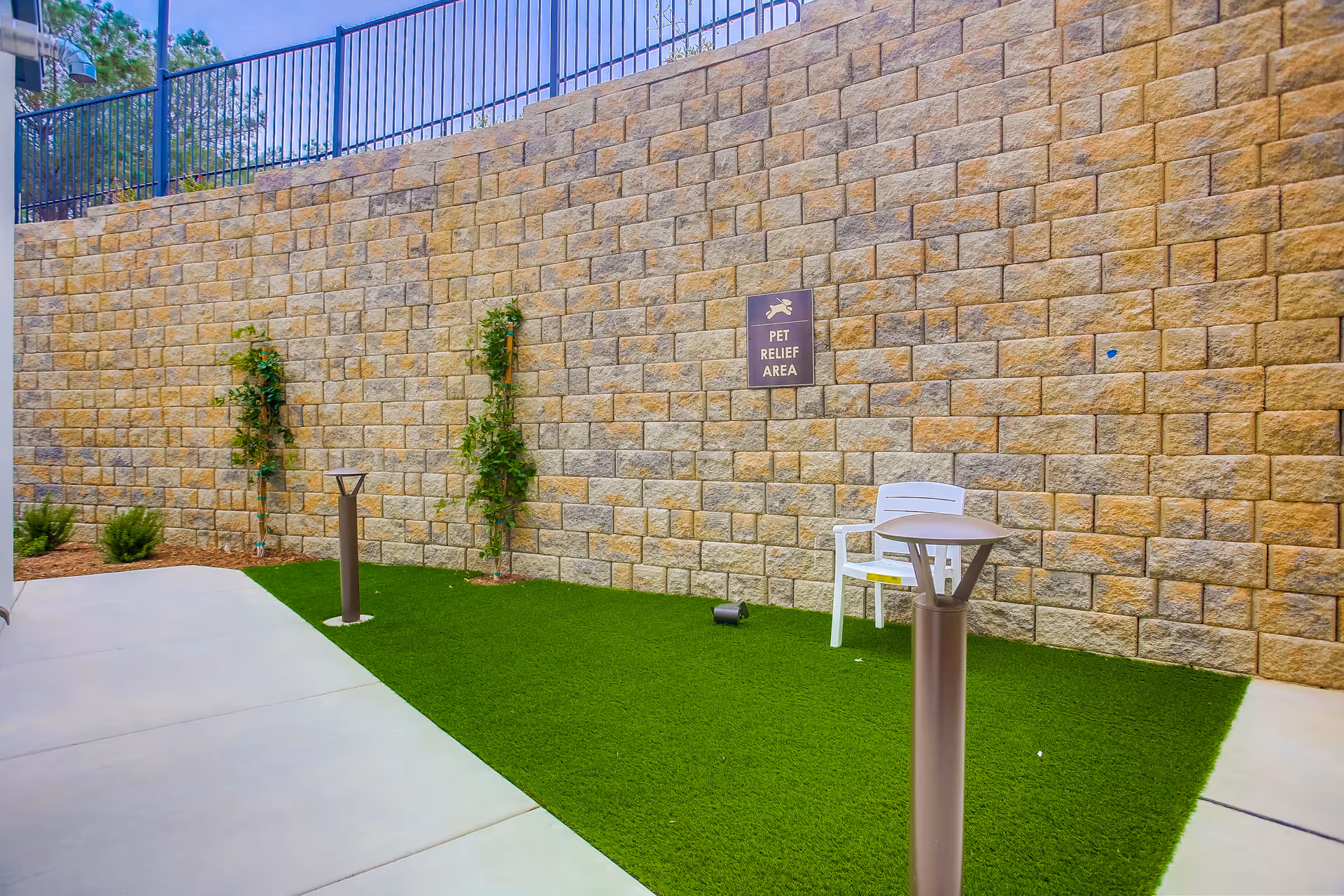 Outdoor pet relief area with artificial grass, a white chair, two small plants growing against a stone wall, and a sign on the wall that reads 'Pet Relief Area'. The area is bordered by concrete walkways and has two modern outdoor lights.