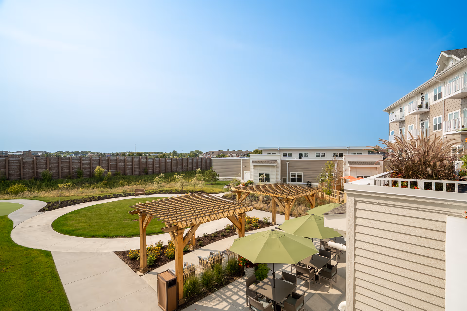 Sunlit outdoor courtyard of a senior living community with wooden pergolas, green umbrella-shaded tables, walking paths, and the adjacent multi-story building.