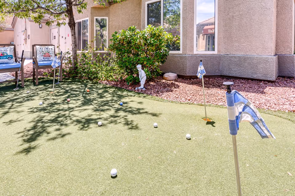 Small outdoor putting green with flags, scattered golf balls, benches and landscaping in front of a stucco building.