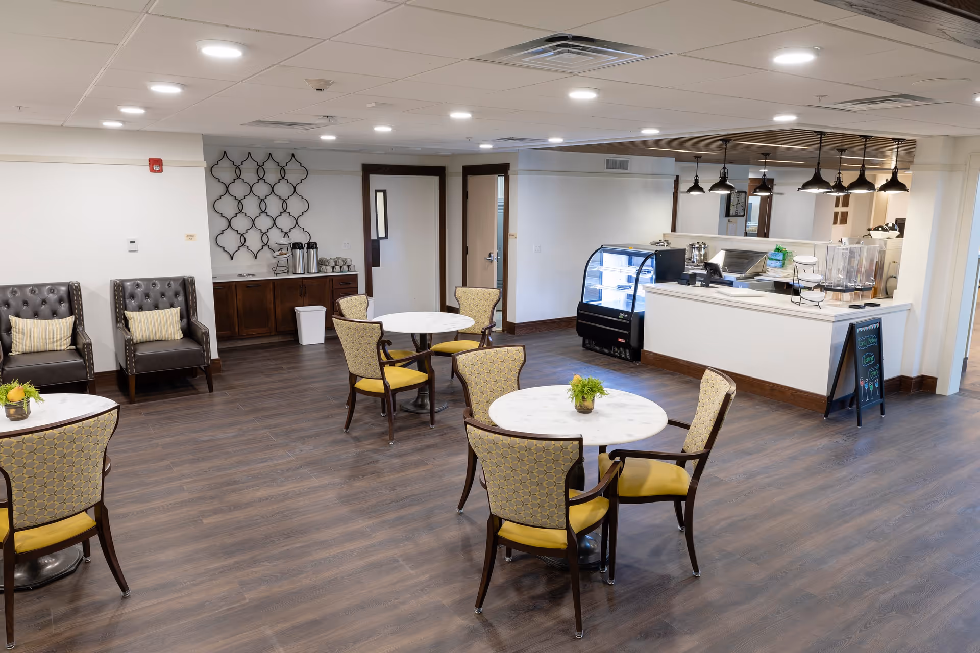 A spacious senior living facility common area with round tables and chairs arranged for seating. There are two leather armchairs with striped cushions against the wall, a small counter with coffee dispensers and cups, and a service counter with a display case and pendant lights overhead. The floor is wood, and the ceiling has recessed lighting.
