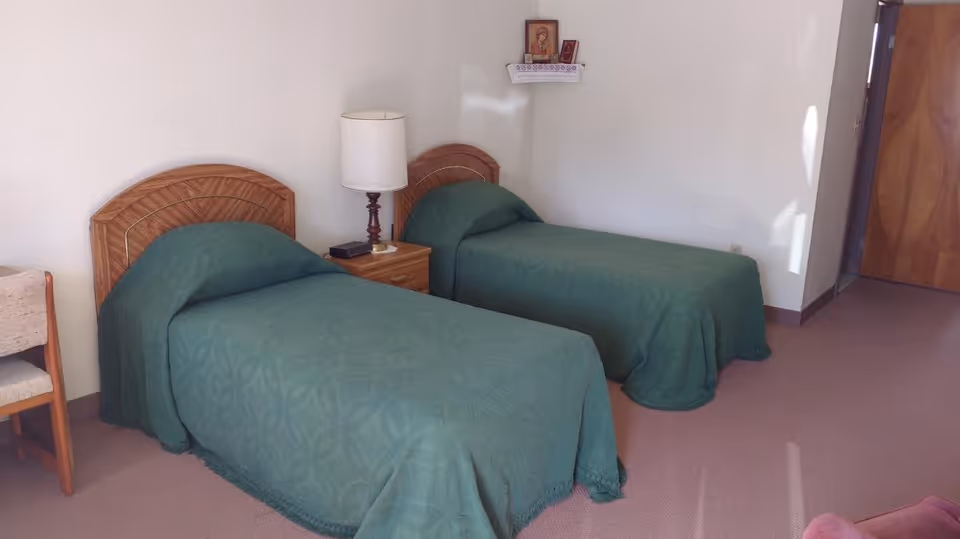Small bedroom with two twin beds covered in green bedspreads, a nightstand with a lamp between them and small religious icons on the wall.