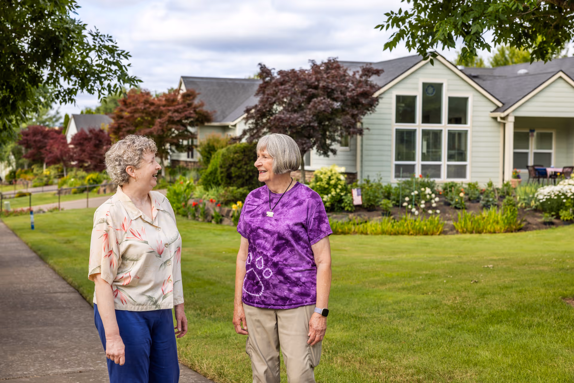 Two elderly women standing and talking on a sidewalk in front of a residential building with well-maintained landscaping, green grass, and trees.