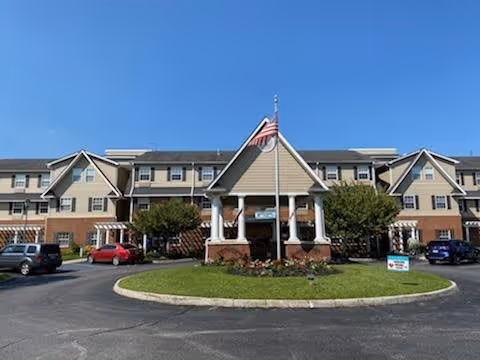 Front exterior view of TerraBella Pheasant Ridge facility showing a large building with multiple windows, a covered entrance with columns, an American flag on a flagpole, several parked cars, and a circular driveway with landscaping in the center.
